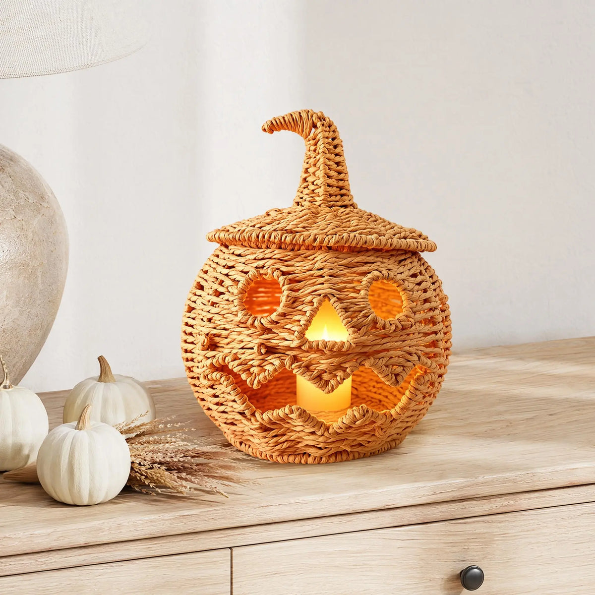 A woven rattan jack-o'-lantern night light with a pointed hat, glowing with warm orange light, displayed on a light wood dresser next to small white pumpkins.