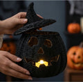 A person holds a black woven pumpkin lantern with a witch hat lid, glowing candles inside, and blurred jack-o'-lanterns in the background.