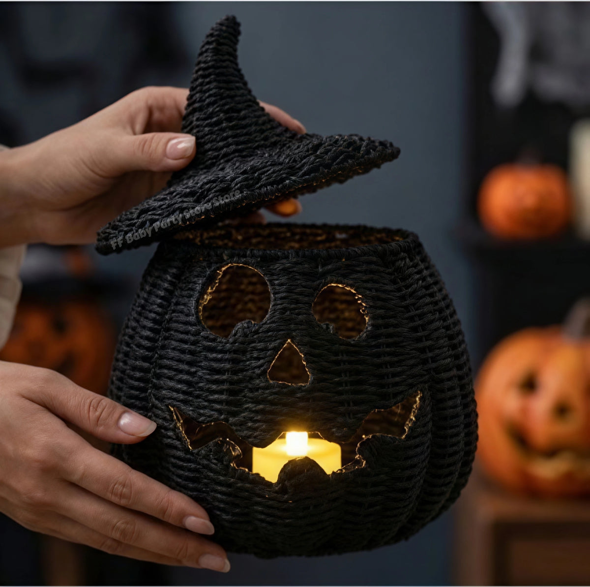 A person holds a black woven pumpkin lantern with a witch hat lid, glowing candles inside, and blurred jack-o'-lanterns in the background.