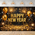 A New Year's Eve party backdrop featuring 'HAPPY NEW YEAR' in glittery gold letters, surrounded by gold and black balloons, fireworks, sparklers, and gold bokeh lights on a black background, with a wooden floor and hanging pendant lights shown above.