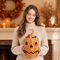 A woman in a beige turtleneck sweater holds a woven pumpkin-shaped candy basket filled with colorful wrapped chocolates in a cozy, warmly lit autumn living room setting.
