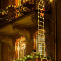 A brick building at night decorated for Christmas, with warm yellow string lights draped around windows and balconies, and a vertical ladder of lights leading up the facade.