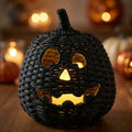 A black woven rattan jack-o'-lantern Halloween decoration with a lit warm yellow interior, shown on a wooden surface with blurred pumpkins and warm lights in the background.
