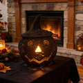 A dark woven pumpkin-shaped lantern with a pointed witch hat sits on a black tablecloth in front of a lit stone fireplace. The lantern has cutout facial features and glows with warm light from within.
