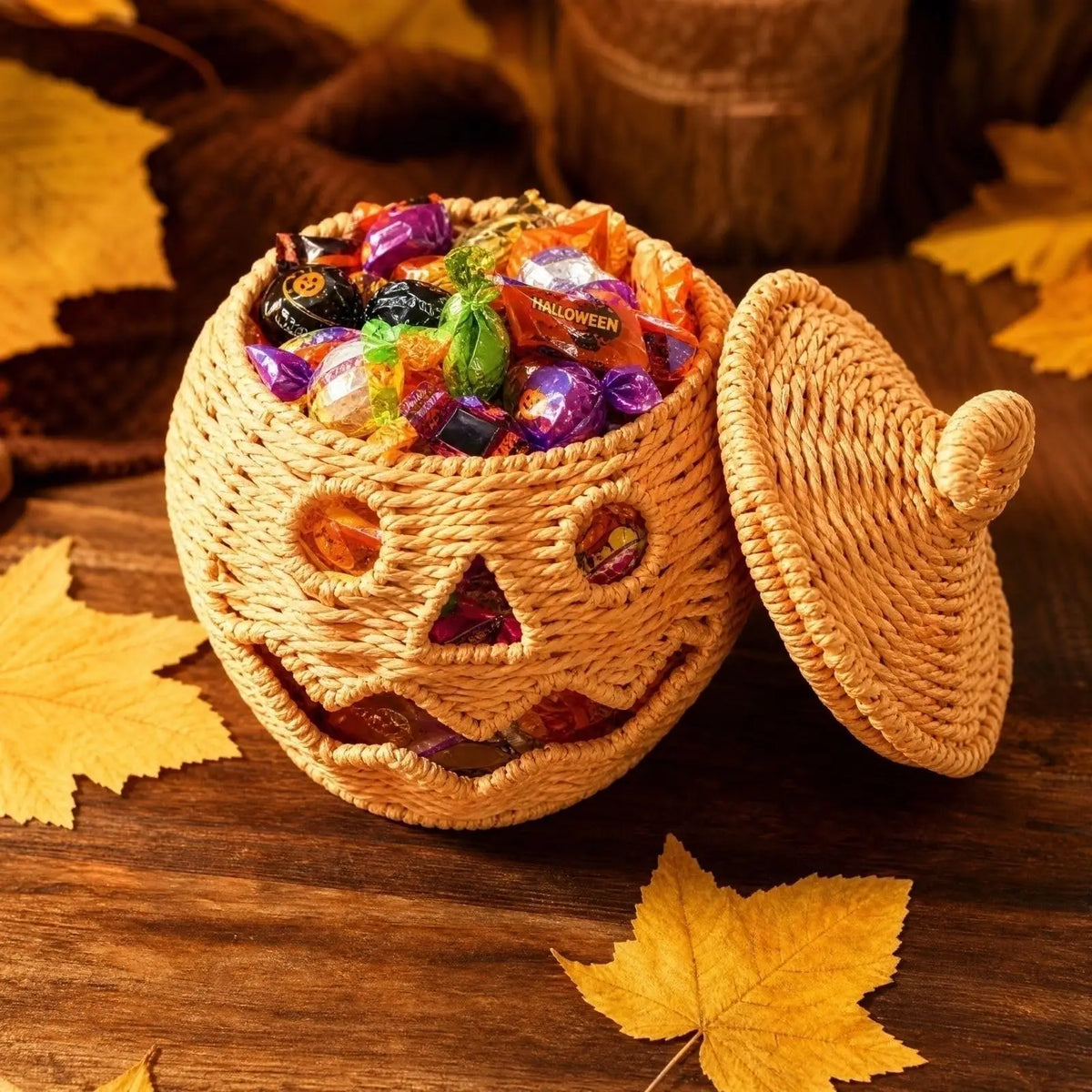 A woven pumpkin-shaped candy container with a lid, filled with assorted Halloween candies, set on a wooden surface with autumn leaves.