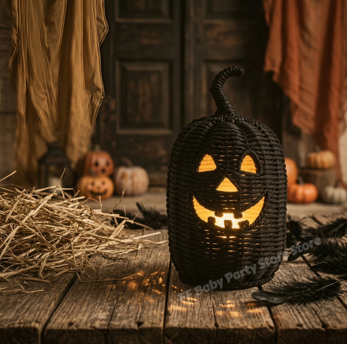 A black woven pumpkin-shaped Halloween lantern with a carved jack-o'-lantern face, glowing with warm yellow light inside, displayed on a rustic wooden surface with hay, dried leaves, and small pumpkins in the background.