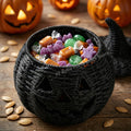A black woven pumpkin-shaped candy bowl filled with assorted Halloween candies, set on a wooden surface with scattered pumpkin seeds and glowing jack-o'-lanterns in the background.