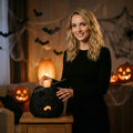 A woman in a black long-sleeve dress stands in a Halloween-decorated room, holding a black woven jack-o'-lantern lantern with a lit candle inside. The background features hanging cobwebs, black bat cutouts, and glowing jack-o'-lanterns on a wooden table.